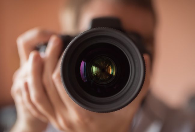 Lens-forward closeup of a camera, held by a photographer. Only the lens and photographer's hands on the camera are in focus.