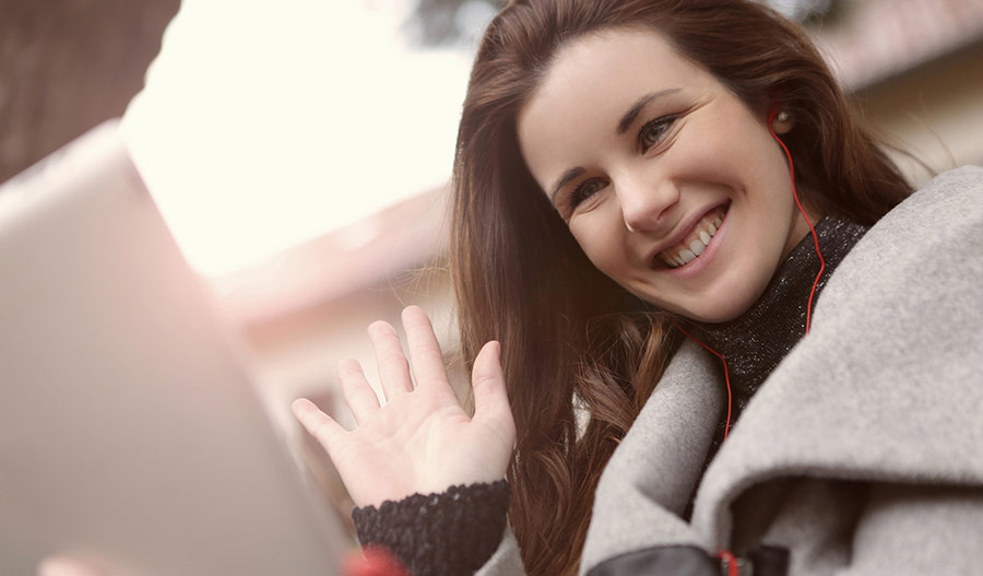 Woman wearing sweater and coat smiling and waving with headphones in her ears