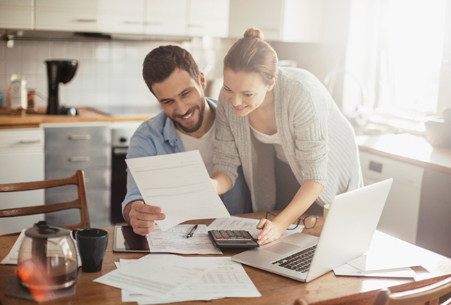 Man sitting at kitchen table holding piece of paper with woman standing next to him pointing at paper. The table also has a laptop, calculator, and several other pieces of paper on it.