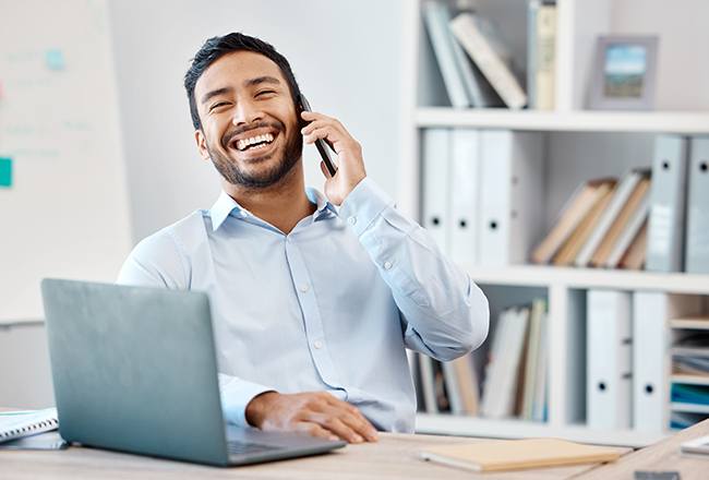 Men leaning back in chair at a desk with a laptop in front of him and holding a cell phone to his ear