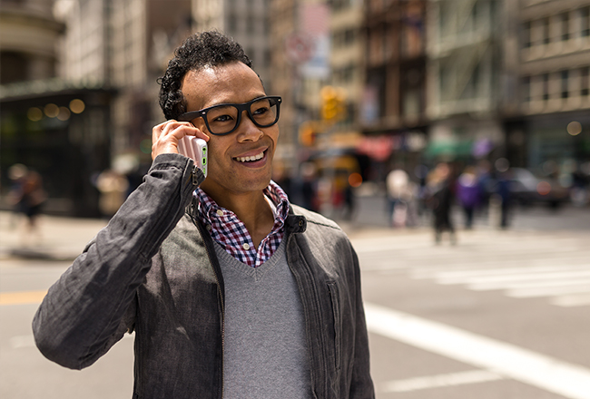 Person with glasses wearing a button-up shirt, sweater, and jacket on sidewalk in city holding cell phone to their ear