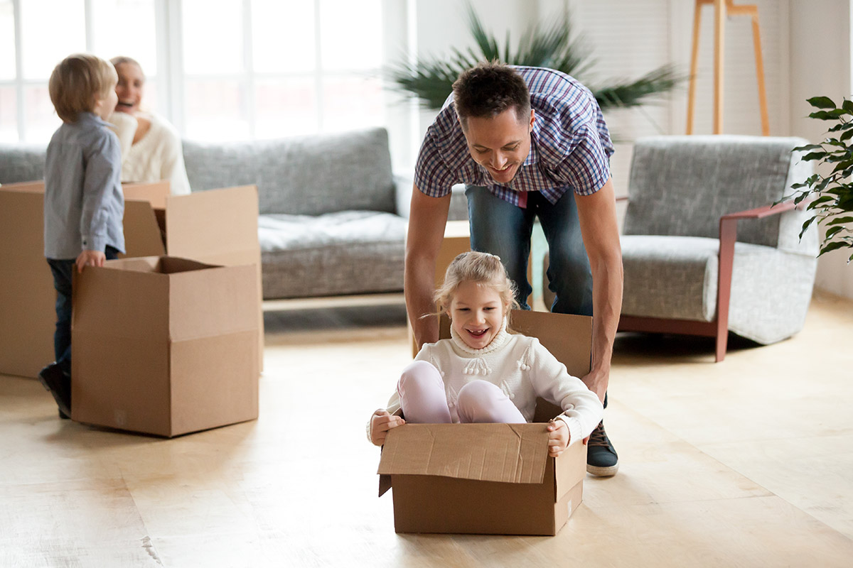 Daddy playing with daughter riding in box on moving day concept, little excited girl enjoying game with father in living room, happy family kids having fun in new house packing preparing to relocate