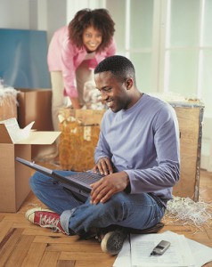 Man searching on laptop while woman packs moving boxes in the background