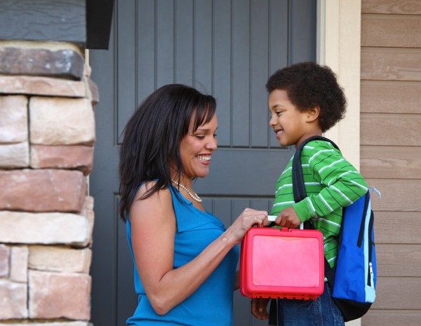 A mother handing a child a lunchbox in front of their home