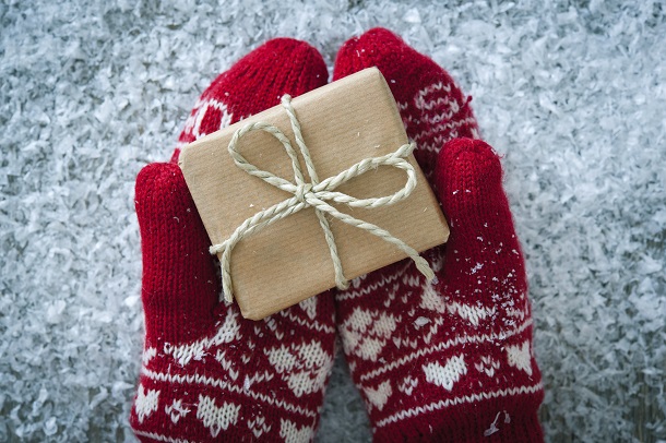 Hands in red and white mittens holding a small gift wrapped in brown paper with a twine bow