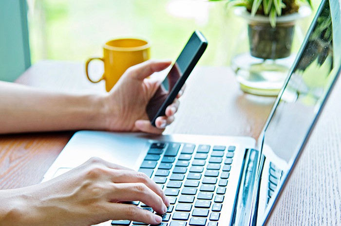 Person typing on laptop while holding cell phone