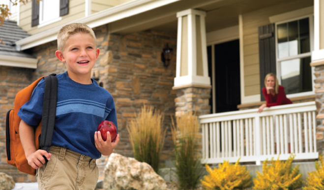 Child with backpack walking in front of home while mother looks on from porch
