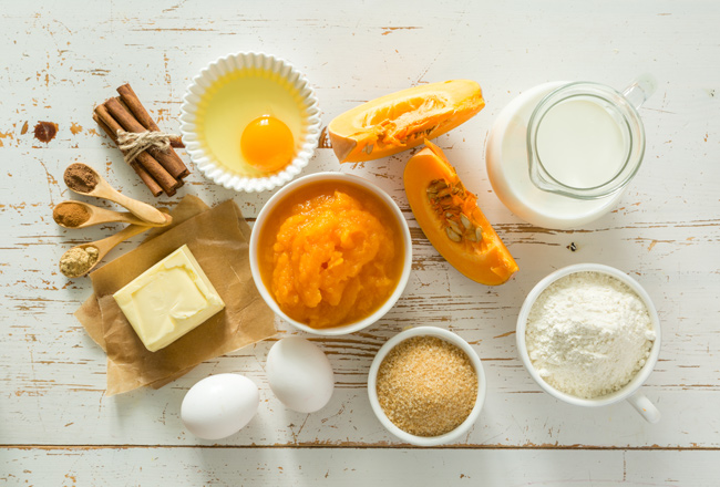 Pumpkin, eggs, cinnamon sticks, brown sugar and other ingredients sitting on white wooden table