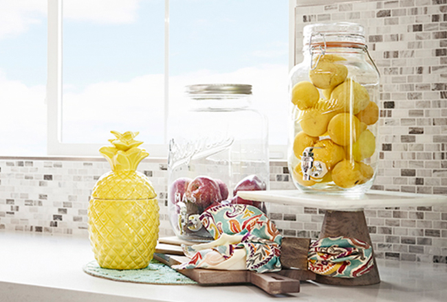 Cutting board, mason jars filled with apples and lemons and a pineapple cookie jar sitting on a kitchen counter