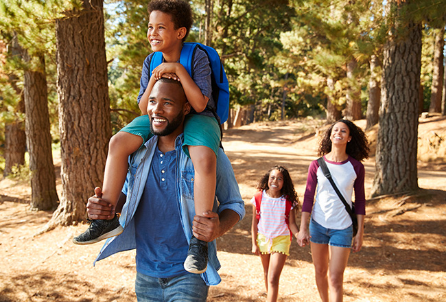 Man walking in woods with son on shoulders followed by woman holding hands with daughter