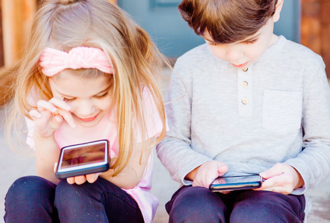 Two children sitting down while looking at devices