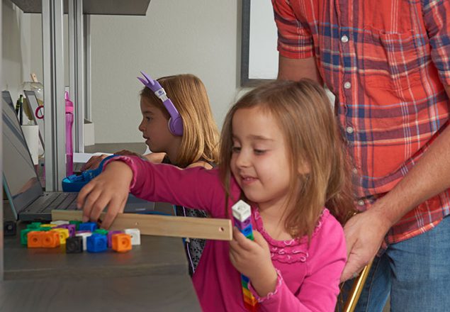 Dad helping girl measure blocks while sister does virtual learning next to her