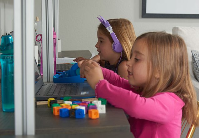 A girl playing with blocks sitting next to her sister doing virtual learning