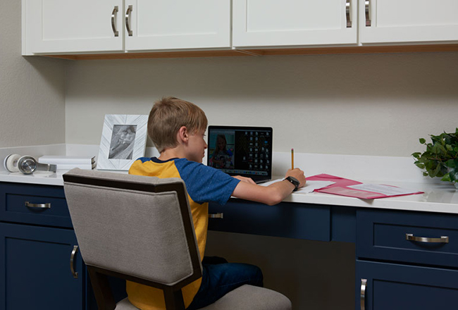 Young boy sitting at built-in desk in front of laptop writing on a piece of paper with a pencil.