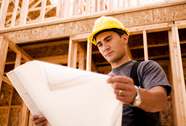 Man wearing a hard hat while reading blueprints in front of a home that is under construction