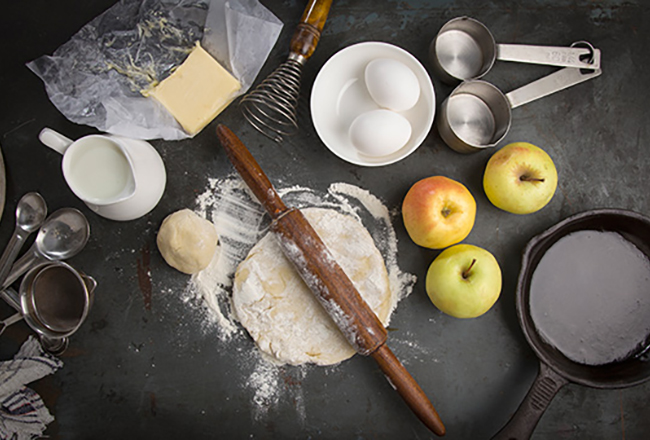 Ingredients and utensils to bake an apple pie, including apples, eggs, measuring cups and a rolling pin