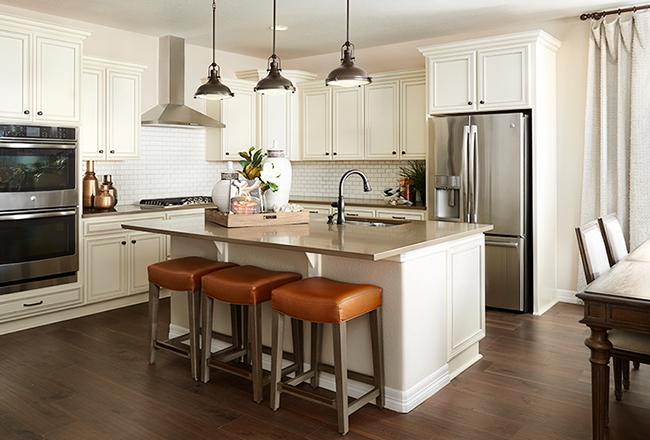 Kitchen with large island with a sink in the center, on the left is a double oven stacked on top of each other, with the stove to the right of it. On the right is the refrigerator, in between the fridge and the stove is counter space and cabinets