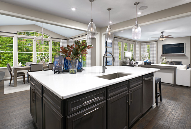 Kitchen with overheard hanging lights, and a large island that includes a sink, behind the island on the left is a very large window and the dining room table. Behind the island on the right is the great hall which includes a couch and a television