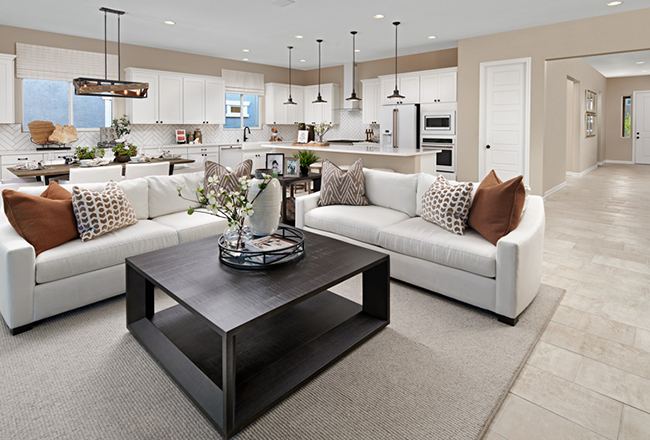 The great room, with the angle showing the kitchen behind and to the right, and the dining room behind and to the left. Two couches forming an 'L' shape, centered between the couches is a large coffee table, The kitchen includes a large island and double ovens, the dining room includes a long table with overhead lighting
