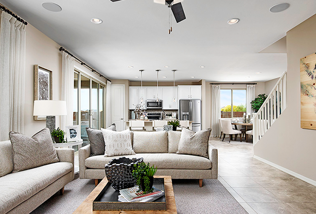 Living room with kitchen shown in the background, on the left side and center of the room there are two couched, that both face a wooden coffee table in the middle of the room. In the corner of the two couches, there is a side table with a lamp