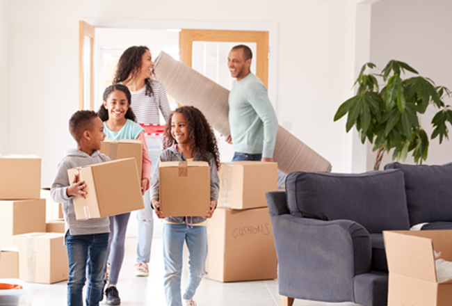 Family with three kids carrying boxes and a rug into a new home with boxes, a couch, and plant