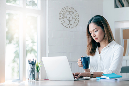 Woman sitting at table while holding coffee mug in right hand and typing on laptop with left hand