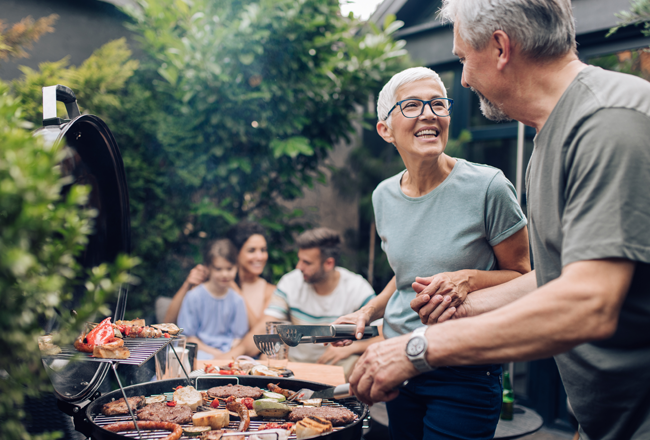Older couple grilling in backyard, with family in the background