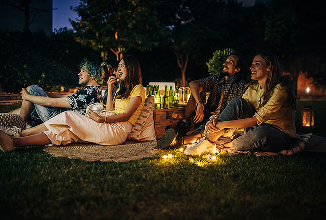 Several people resting on blankets and pillows outdoors at night, apparently watching a movie that's off camera