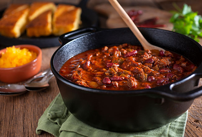 Cast-iron pot of chili with a plate of cornbread and dish of grated cheese in the background