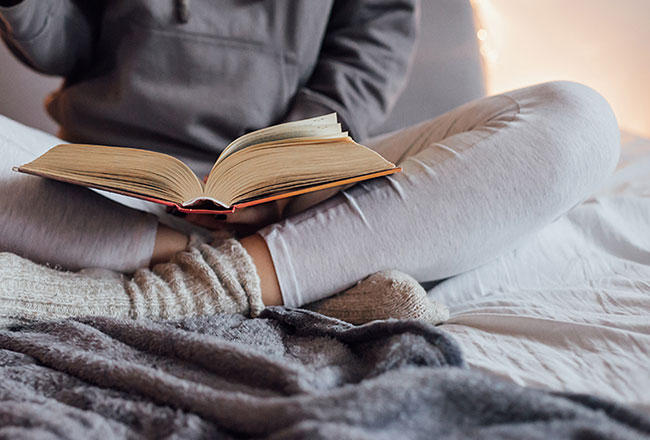 A person in warm clothing, sitting cross-legged and reading a book