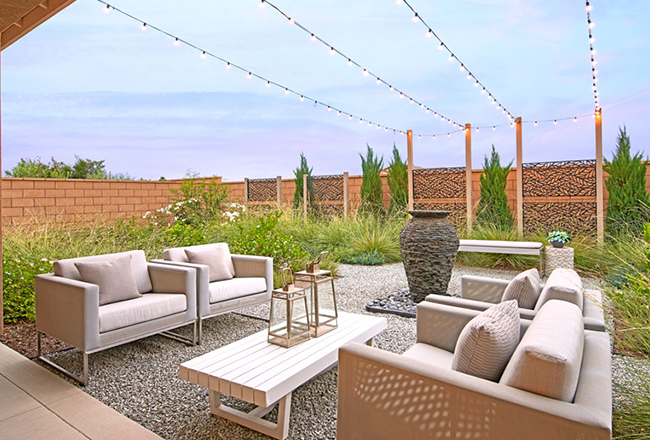 Backyard with lounge chairs below string lights, surrounded by plants and grasses