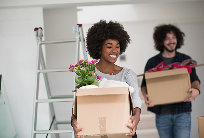 Woman carrying a cardboard box with a plant sticking out the top, and a man behind her carrying another box