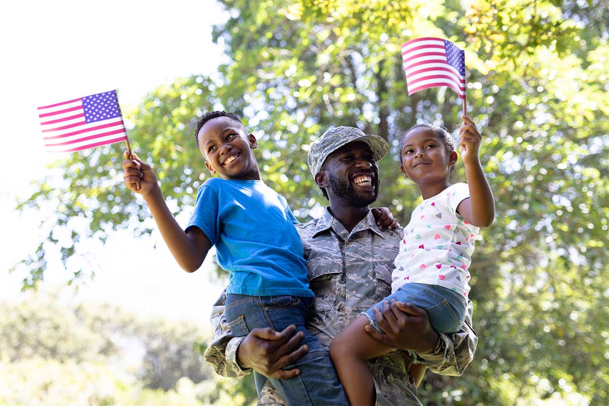 Man in military garb holding two children who are holding up American flags