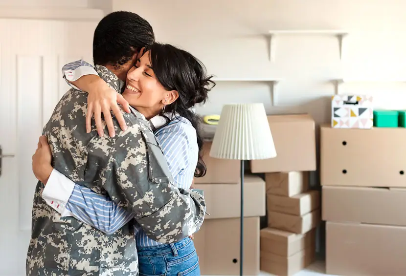 A woman hugging someone in a military uniform, with moving boxes in the background