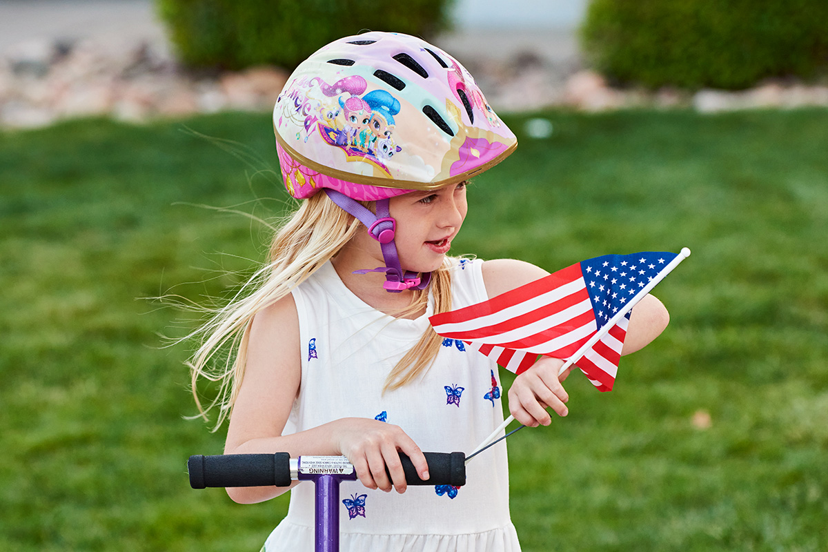 young girl scootering, wearing a helmet and holding USA flag