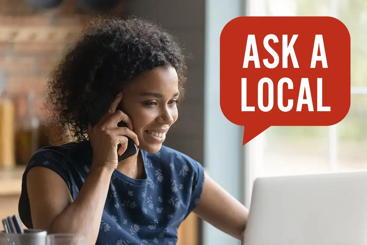 Woman sitting at table looking at laptop while holding cell phone to ear and smiling, with 'Ask a Local' blurb overtop