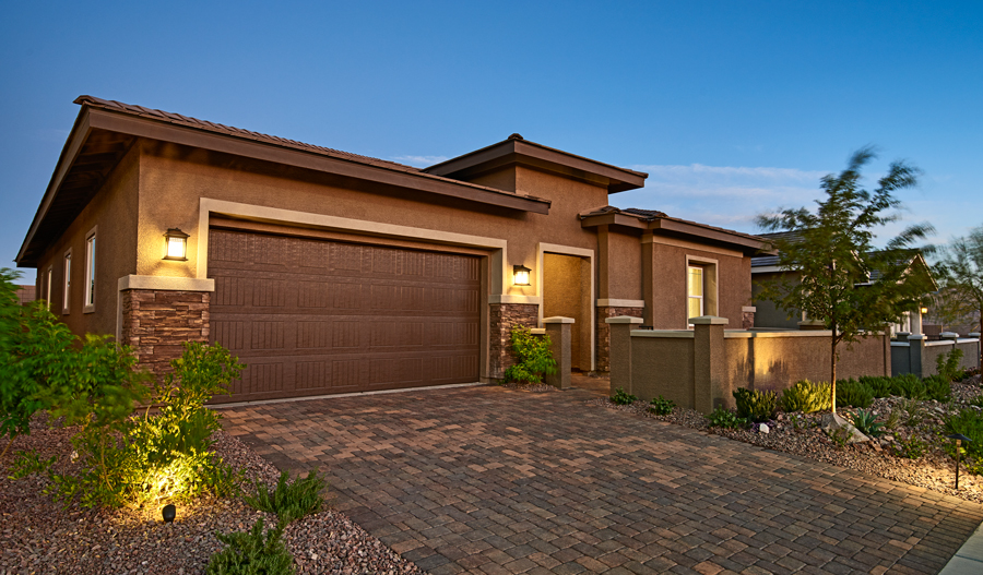 The exterior of a ranch style home, with a stone driveway and two car garage. Rocks and plants landscape the front yard, and there is a wall guarding the covered entryway from the street