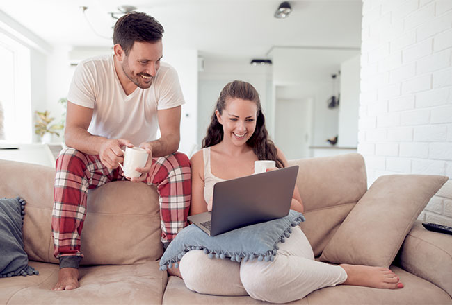 Woman sitting on couch with pillow and laptop on her lap, while man sits behind her on the top of the couch looking