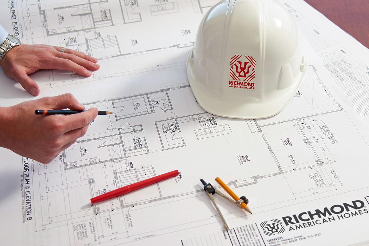Architectural drawings with hardhat and red marker on top and a hand hovering over with pencil