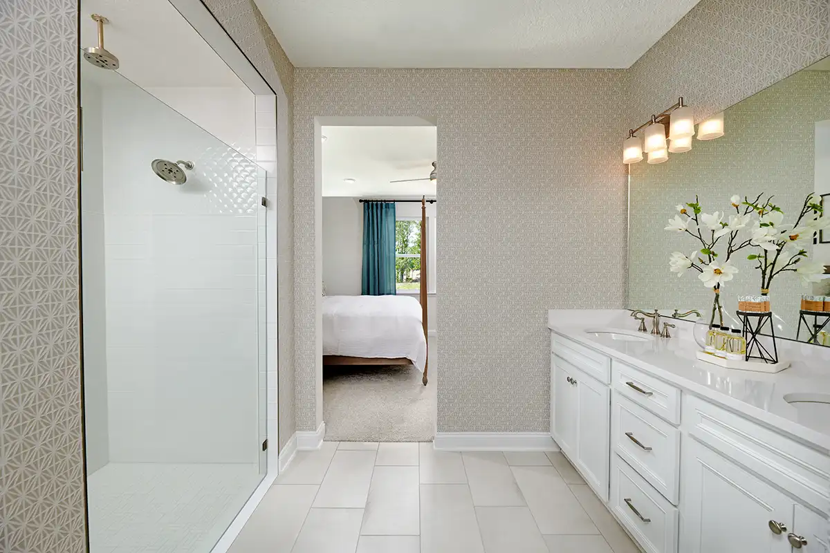 Bathroom with patterned wallpaper, two-sink vanity below large mirror, shower stall with two showerheads, and doorway to bedroom