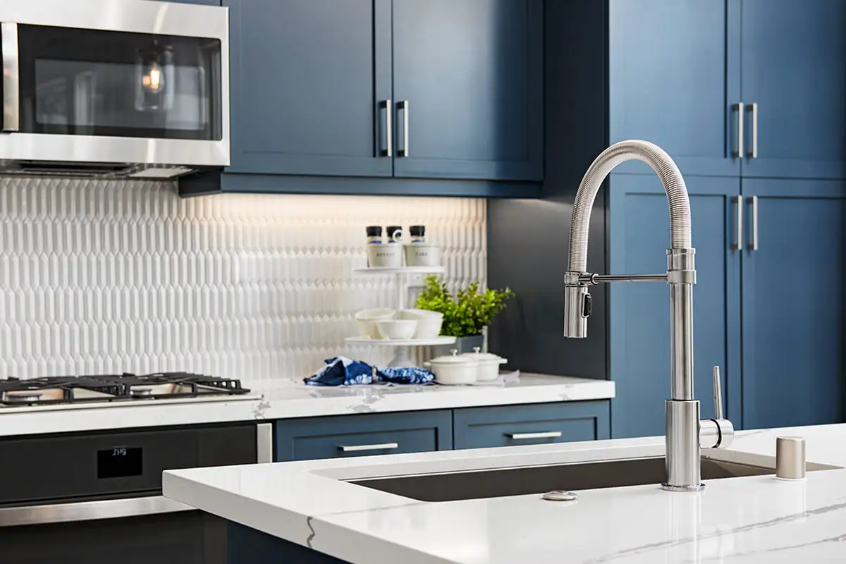 Kitchen sink on island with blue cabinets, this picket backsplash, gas stove, and microwave in the background