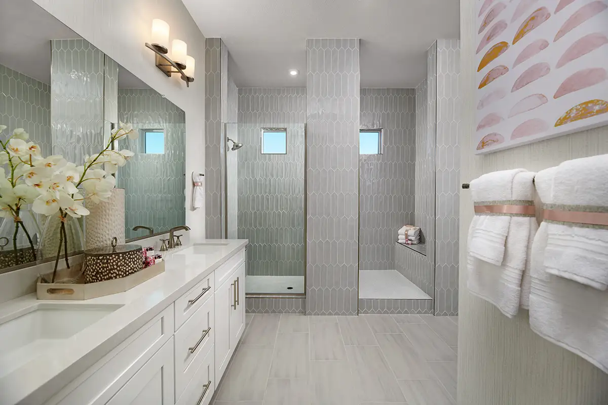 Bathroom with large light double sink vanity with gold hardware and step up into large shower stall with two small windows and seat
