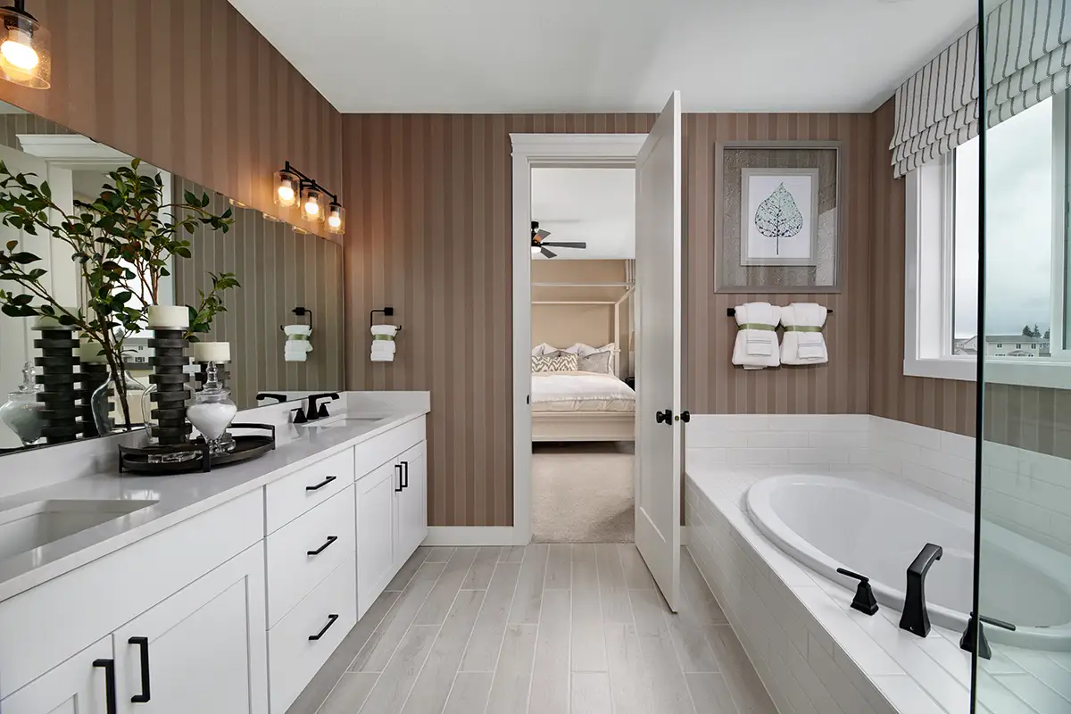 Large bathroom with double sinks, bathtub, striped wallpaper, and an open door leading into the owner's bedroom