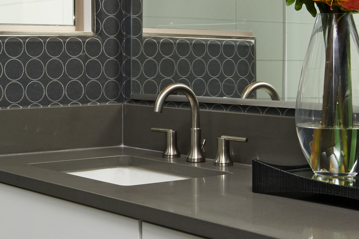 Bathroom sink with dark countertops, silver hardware, and a circular patterned wallpaper below a window
